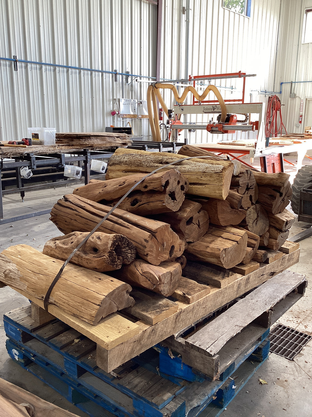 A pile of rough-sanded, debarked Osage Orange wood table legs stacked on a wooden pallet in a warehouse setting.