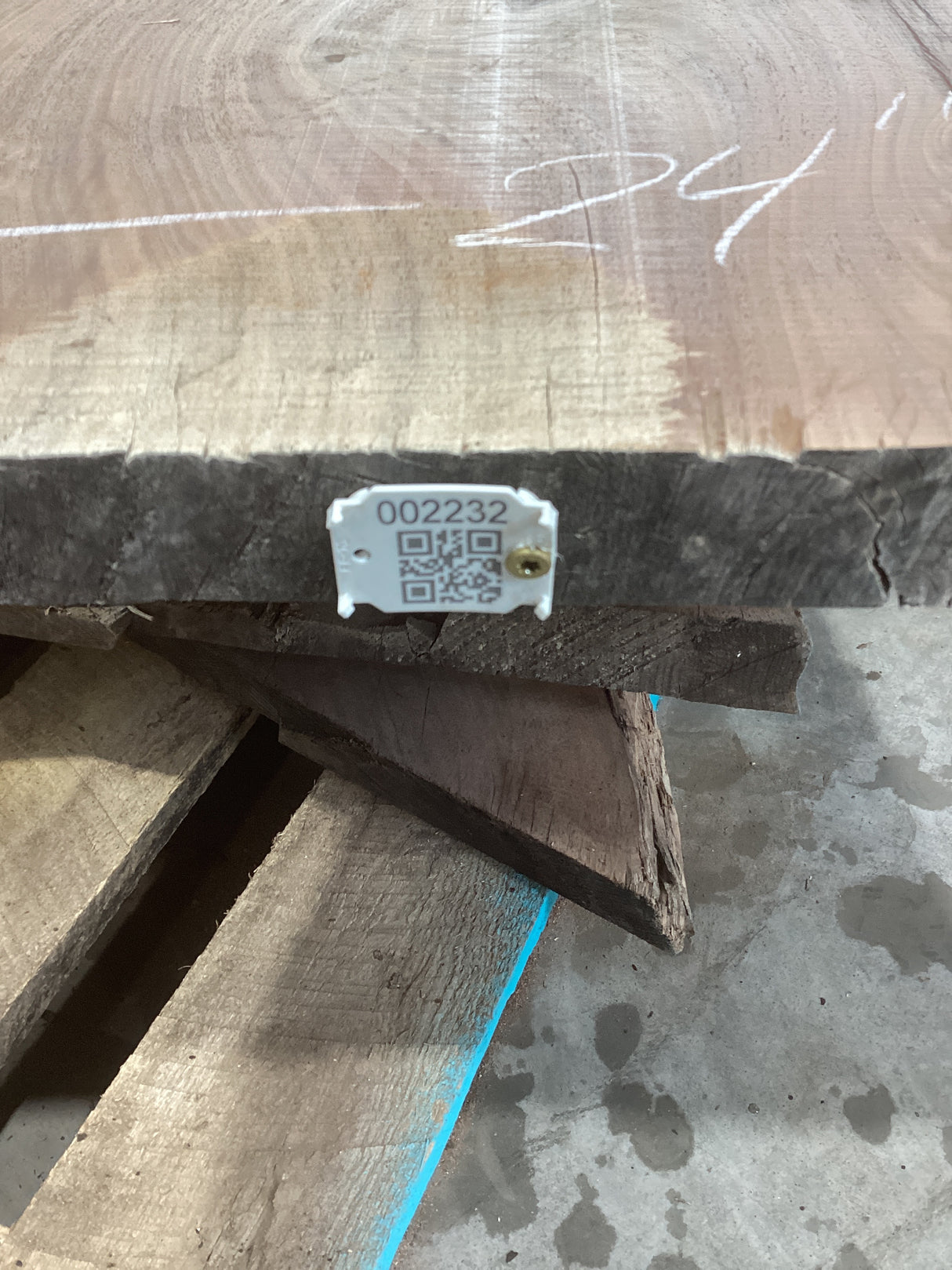 A walnut wood slab with a visible wood grain and a QR code tag attached to it, resting on a pile of wood.