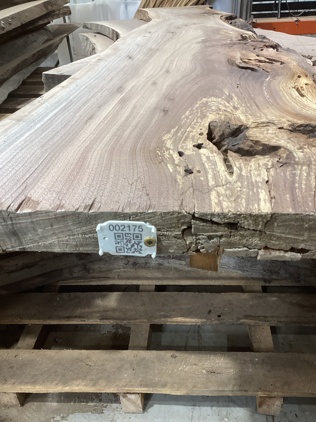 A walnut wood slab with natural edges and a visible QR label attached to it, placed on a wooden pallet.