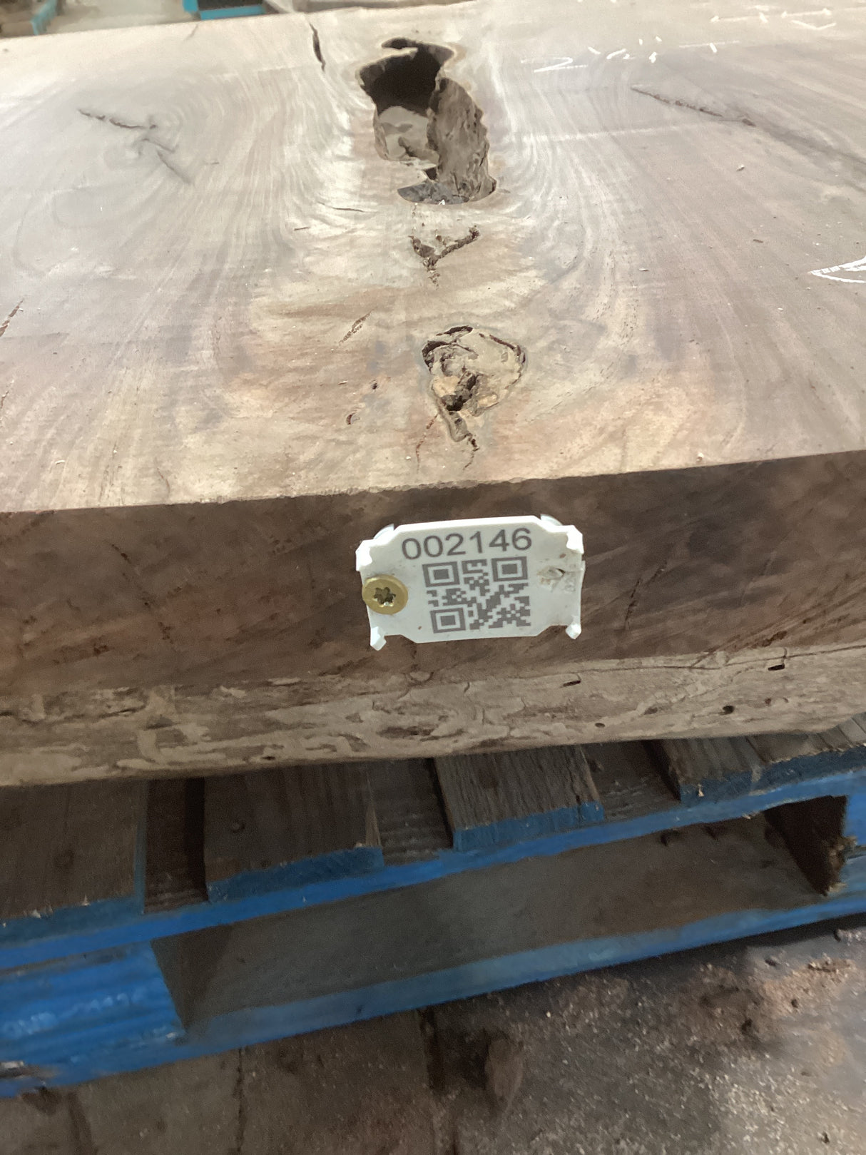 A walnut slab with a distinctive grain pattern and a  QR tag attached to it, resting on a blue pallet.