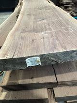 A walnut slab with a natural edge, laid flat on a surface, with a tag attached to one end displaying a barcode and product information.