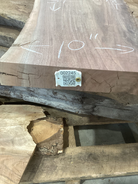 A walnut wood slab placed on a wooden rack, with a QR tag attached to it showing a barcode and product information.
