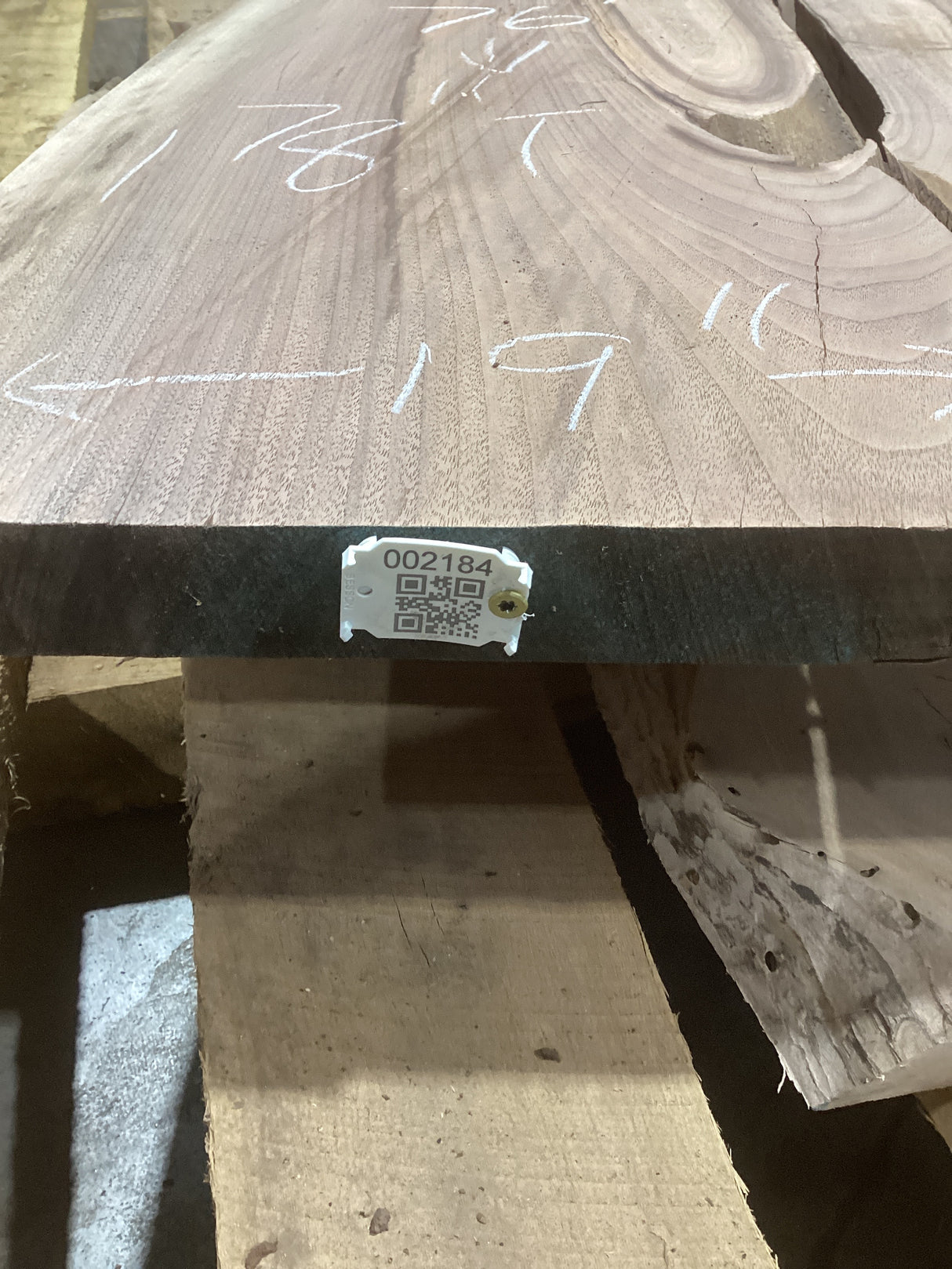 A walnut wood slab with visible natural patterns and a QR label on the edge. The slab is placed on a wooden support structure, and there are measurements marked on the surface.