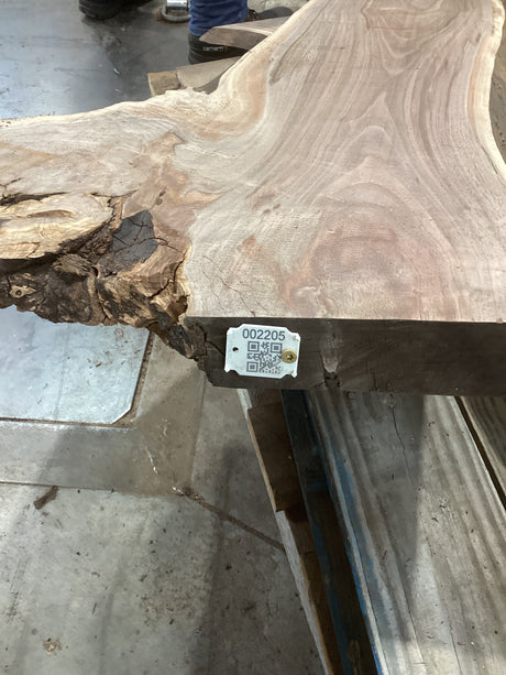 A large walnut slab with visible wood grain patterns, positioned on a metal table frame, with a QR tag attached to one end.