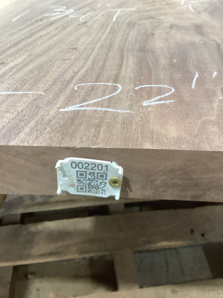 A walnut wood slab with a visible label that has a QR code and the number 002201 written on it. The slab is placed on a stack of wood in a workshop setting.
