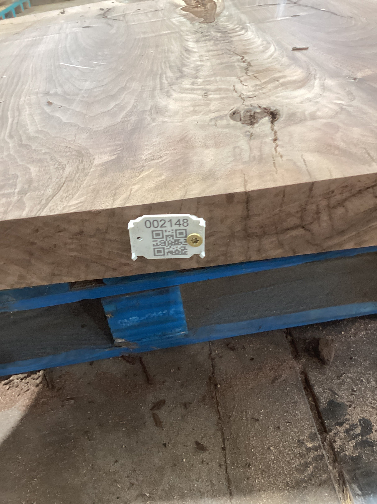 A walnut wood slab with a visible grain pattern and a QR tag attached to it, placed on a blue surface with a concrete floor in the background.