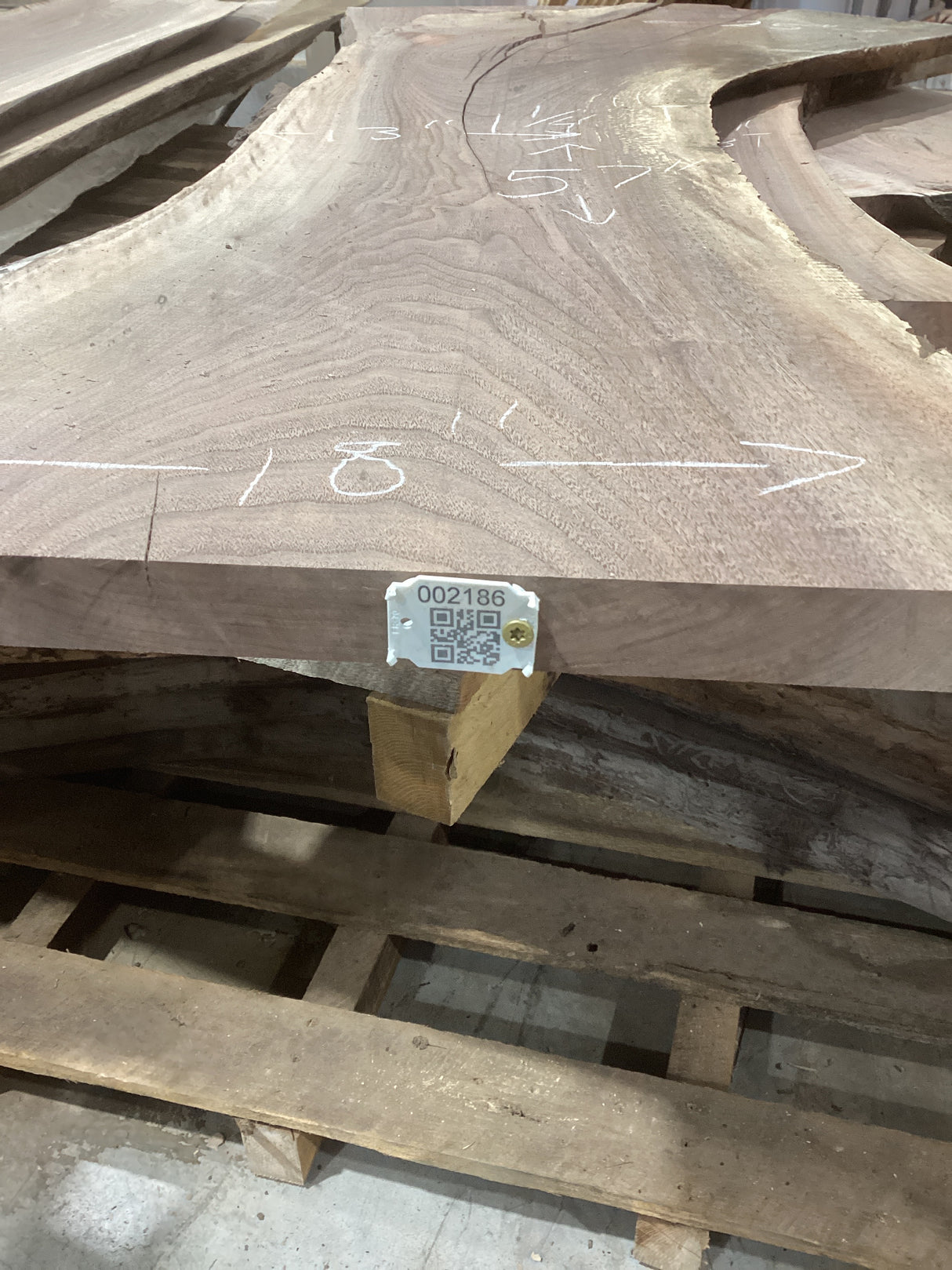 A walnut slab with natural color variations and a visible QR label attached to it, placed on a wooden pallet.