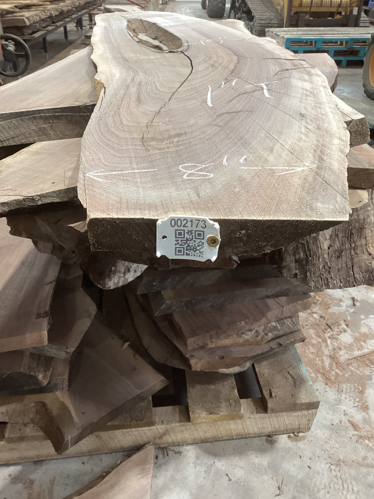 A walnut slab with a natural 'lake' in the center, showing the wood grain and a visible missing sliver, placed on a stack of other wood slabs. QR label attached to the edge.