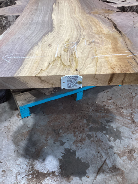A walnut slab with unique contrasting color patterns placed on a blue support structure, with a QR label attached to the edge.