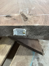 A walnut slab with a live edge featuring a natural wood grain pattern and an ingrown bark causing a crotch slab separation. A label with a QR code is attached to the slab.
